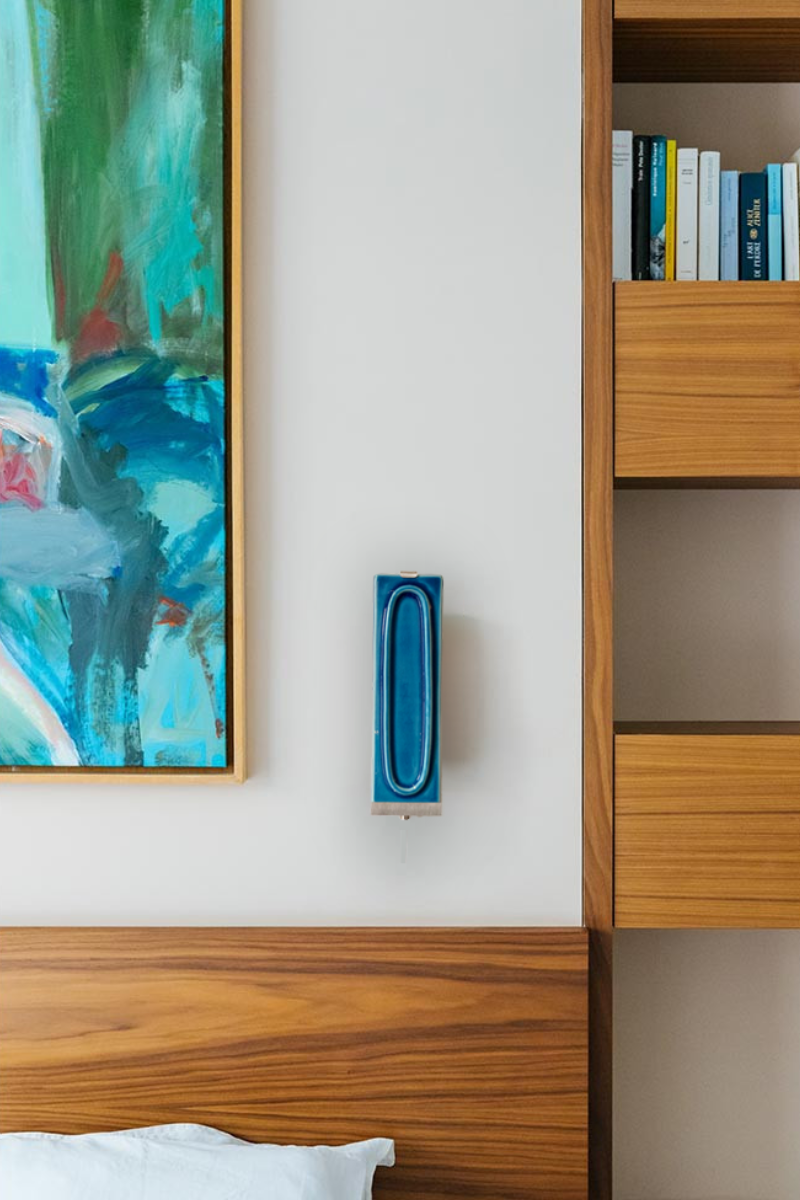 Applique arty bleu dans une ambiance de chambre, avec tableau mural et rangements en bois. L’applique Arty, imaginée par Coline Leleu et Sébastien Carette, porte bien son nom : elle apporte une touche artistique discrète qui habille le mur avec légèreté. Chaque carreau de céramique est façonné à la main au Portugal, et révèle ses nuances et textures uniques, témoignant du geste de l’artisan.