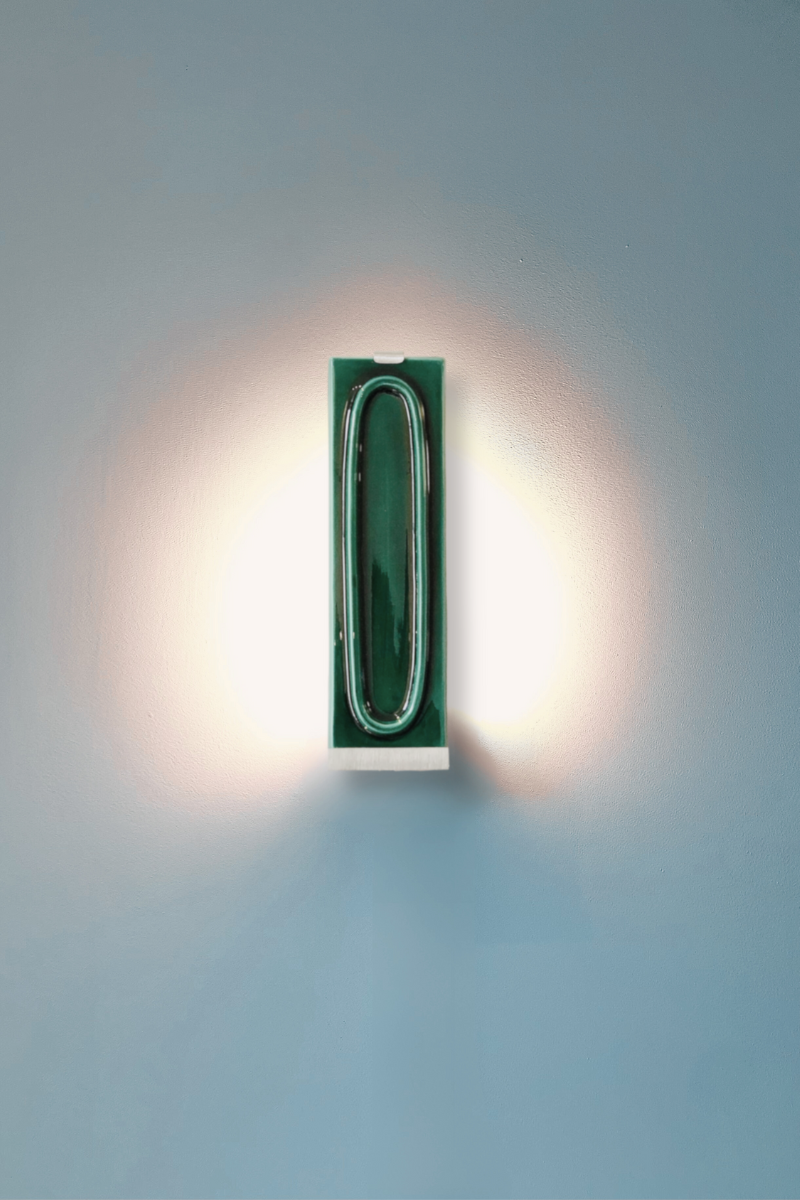Zoom sur l'applique arty verde éclairée.. L’applique Arty, imaginée par Coline Leleu et Sébastien Carette, porte bien son nom : elle apporte une touche artistique discrète qui habille le mur avec légèreté. Chaque carreau de céramique est façonné à la main au Portugal, et révèle ses nuances et textures uniques, témoignant du geste de l’artisan.