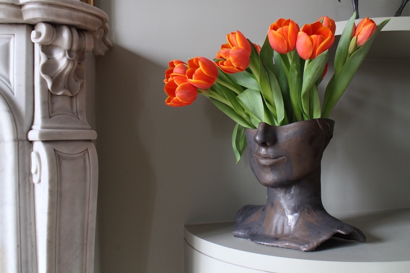 Vase Tête de Bronze by Thalia Dalecky, in glazed orange stoneware: a hand-sculpted face in high relief reveals golden reflections in the light, strength and delicacy fused between ceramic and bronze.