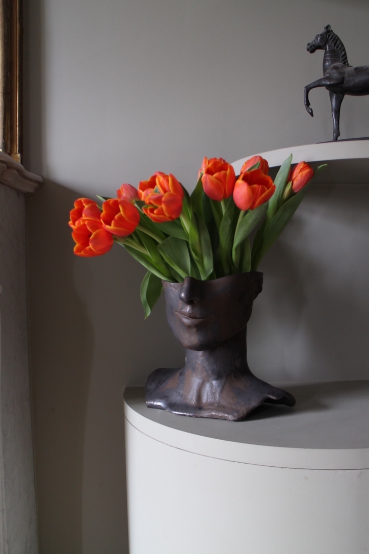 Le vase Tête de Bronze est réalisé par l'artiste Thalia Dalecky avec du grès orange. Sa surface patinée dévoile un visage sculpté dans un grès émaillé noir métallisé.&nbsp; À la lumière, des reflets dorés jouent sur ses contours, ajoutant une touche de mystère à cette pièce d'art captivante, où le bronze robuste rencontre la délicatesse du grès.