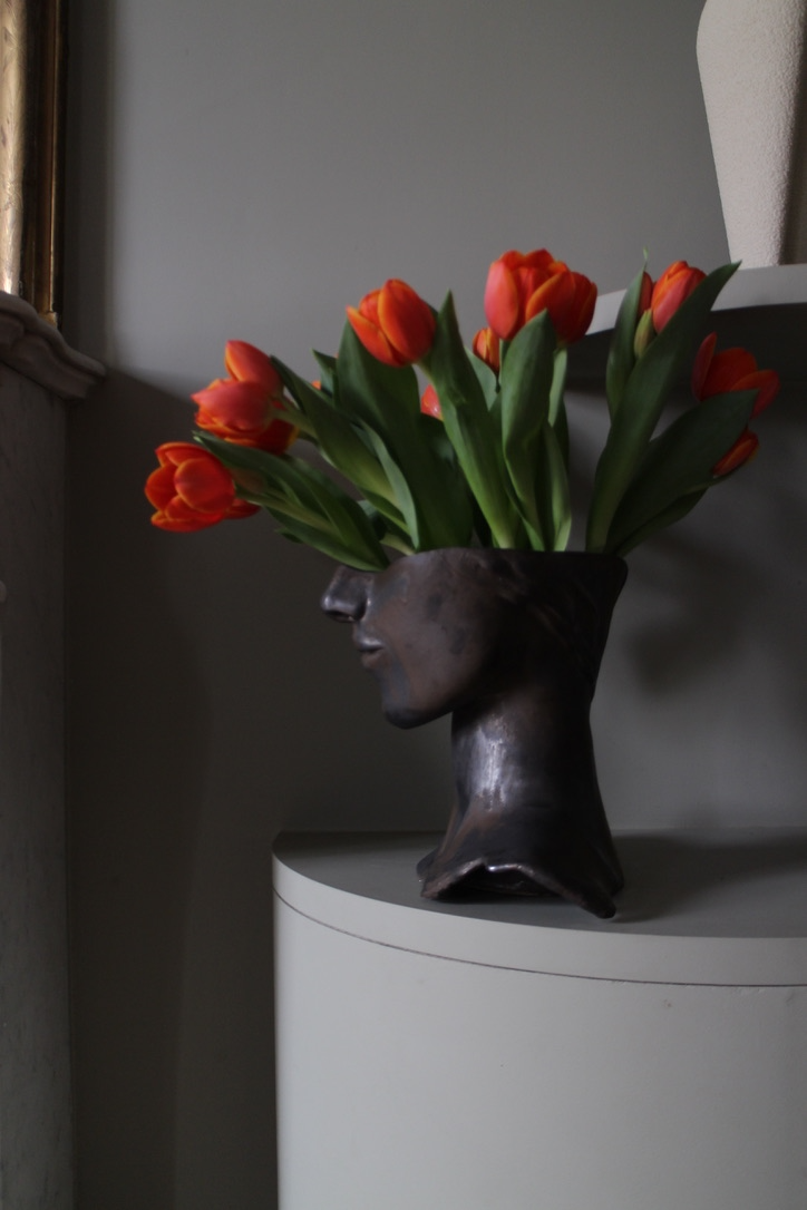 Vase Tête de Bronze by Thalia Dalecky, in glazed orange stoneware: a hand-sculpted face in high relief reveals golden reflections in the light, strength and delicacy fused between ceramic and bronze.