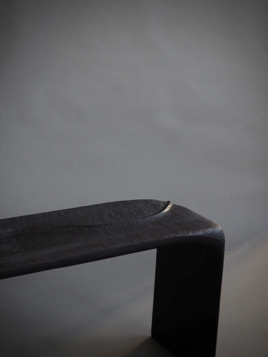 Banc en bois sculpté et brûlé, pensé et façonné à la main par Maxime Perrolle comme un paysage intime. Sa silhouette fluide semble avoir été modelée par le temps, creusée, polie, habitée par le geste. La surface, volontairement texturée, capte la lumière et révèle un jeu subtil d’ombres et de reliefs, invitant autant au regard qu’au toucher.

À la frontière entre mobilier et sculpture, ce banc impose une présence calme et puissante.