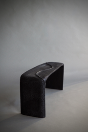 Banc en bois sculpté et brûlé, pensé et façonné à la main par Maxime Perrolle comme un paysage intime. Sa silhouette fluide semble avoir été modelée par le temps, creusée, polie, habitée par le geste. La surface, volontairement texturée, capte la lumière et révèle un jeu subtil d’ombres et de reliefs, invitant autant au regard qu’au toucher.

À la frontière entre mobilier et sculpture, ce banc impose une présence calme et puissante.