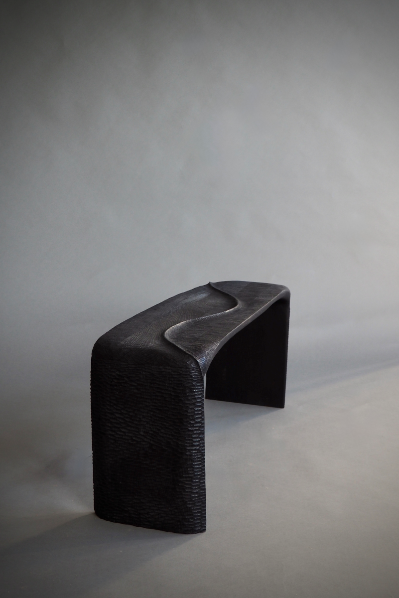 Banc en bois sculpté et brûlé, pensé et façonné à la main par Maxime Perrolle comme un paysage intime. Sa silhouette fluide semble avoir été modelée par le temps, creusée, polie, habitée par le geste. La surface, volontairement texturée, capte la lumière et révèle un jeu subtil d’ombres et de reliefs, invitant autant au regard qu’au toucher.

À la frontière entre mobilier et sculpture, ce banc impose une présence calme et puissante.