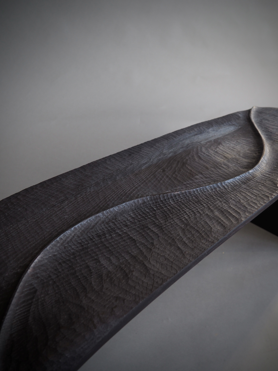 Banc en bois sculpté et brûlé, pensé et façonné à la main par Maxime Perrolle comme un paysage intime. Sa silhouette fluide semble avoir été modelée par le temps, creusée, polie, habitée par le geste. La surface, volontairement texturée, capte la lumière et révèle un jeu subtil d’ombres et de reliefs, invitant autant au regard qu’au toucher.

À la frontière entre mobilier et sculpture, ce banc impose une présence calme et puissante.