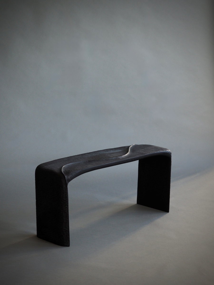 Banc en bois sculpté et brûlé, pensé et façonné à la main par Maxime Perrolle comme un paysage intime. Sa silhouette fluide semble avoir été modelée par le temps, creusée, polie, habitée par le geste. La surface, volontairement texturée, capte la lumière et révèle un jeu subtil d’ombres et de reliefs, invitant autant au regard qu’au toucher.

À la frontière entre mobilier et sculpture, ce banc impose une présence calme et puissante.