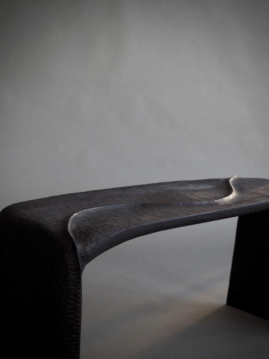 Banc en bois sculpté et brûlé, pensé et façonné à la main par Maxime Perrolle comme un paysage intime. Sa silhouette fluide semble avoir été modelée par le temps, creusée, polie, habitée par le geste. La surface, volontairement texturée, capte la lumière et révèle un jeu subtil d’ombres et de reliefs, invitant autant au regard qu’au toucher.

À la frontière entre mobilier et sculpture, ce banc impose une présence calme et puissante.