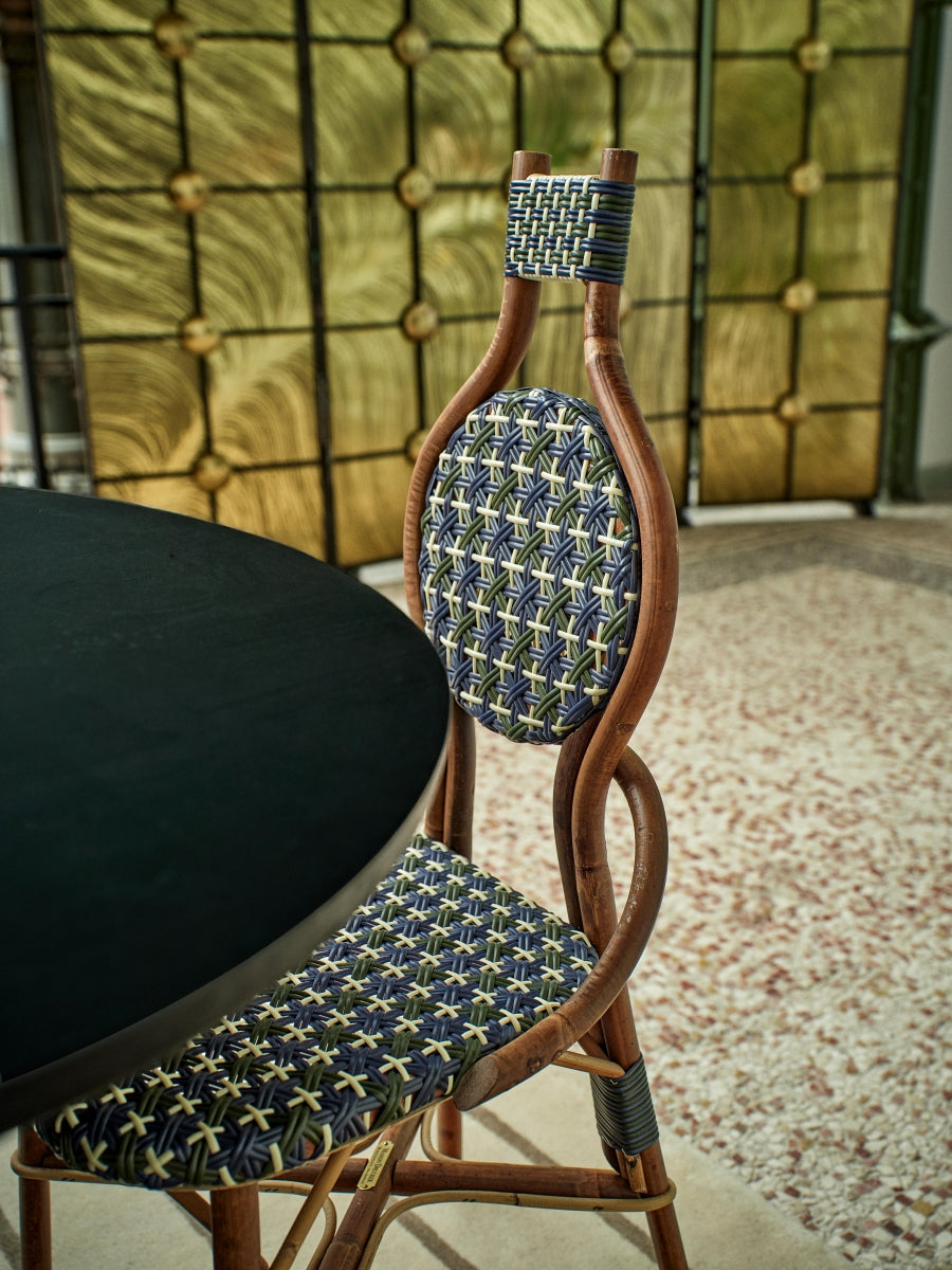 La chaise Odette fait la part belle au rotin. Le Second Empire a vu se développer le métier de rotinier et a été l’âge d’or de ce matériau. Le rotin est une tige de palmier importée des forêts tropicales asiatiques à l’époque coloniale. La chaise de bistro en rotin symbolise à elle seule les terrasses parisiennes et la gaieté de la capitale française. La forme florale et l’utilisation de matières végétales pour le design de la chaise Odette est une référence au catleya.