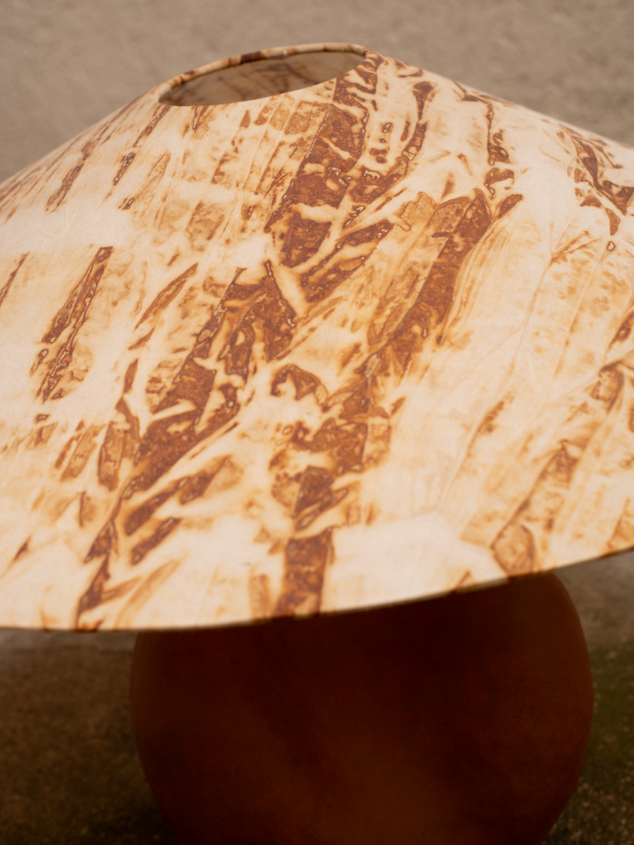 Lampe à poser conçue et fabriquée par Marguerite Porche en collaboration avec Sandrina Rocha, artiste textile. Pied en grès rouge brut de forme ronde, enveloppante, surmonté d’un large abat-jour conique en soie teint naturellement à l’aide de végétaux.