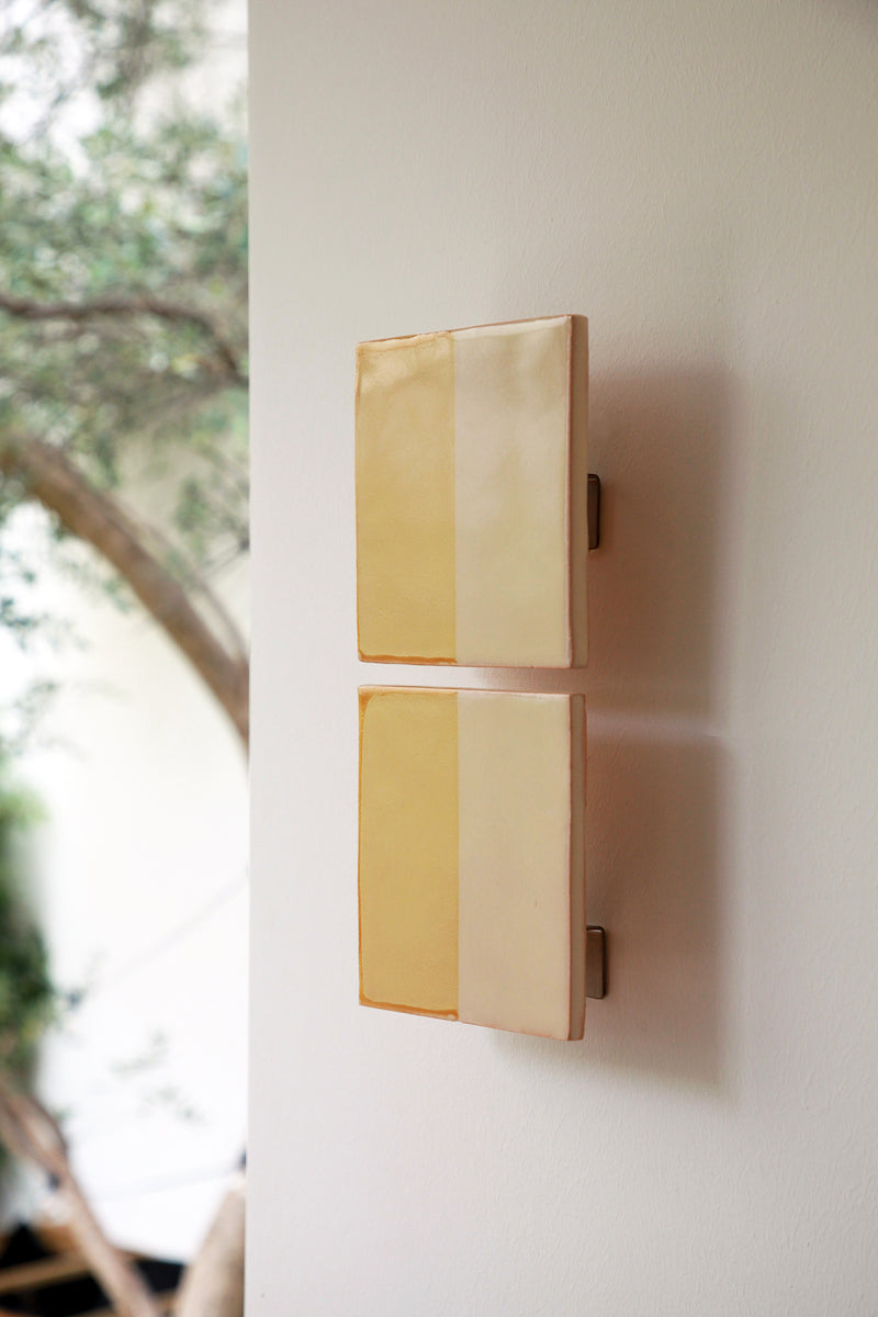 Les Tiles sont des appliques murales conçues par Violaine d'Harcourt en collaboration avec la designer hollandaise Marianne Smink. Utilisant le procédé de la sérigraphie, l'applique Tiles G est ainsi colorée d'un jaune et d'un beige représentant la céramique brute. L'ampoule, cachée derrière un carreau, diffuse un éclairage indirect, créant un effet de clair-obscur et un éclairage très doux.